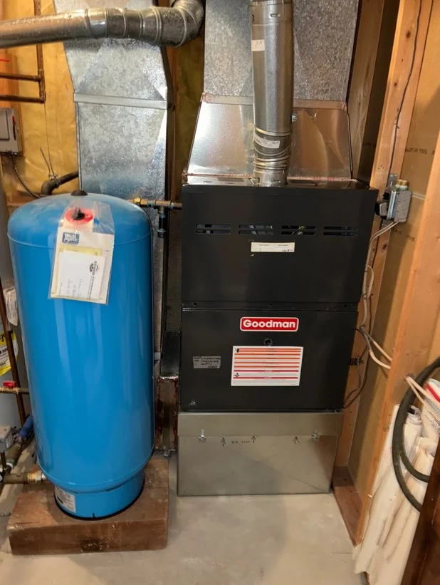 Goodman furnace with blue pressure tank, metal ductwork, and piping in basement utility room